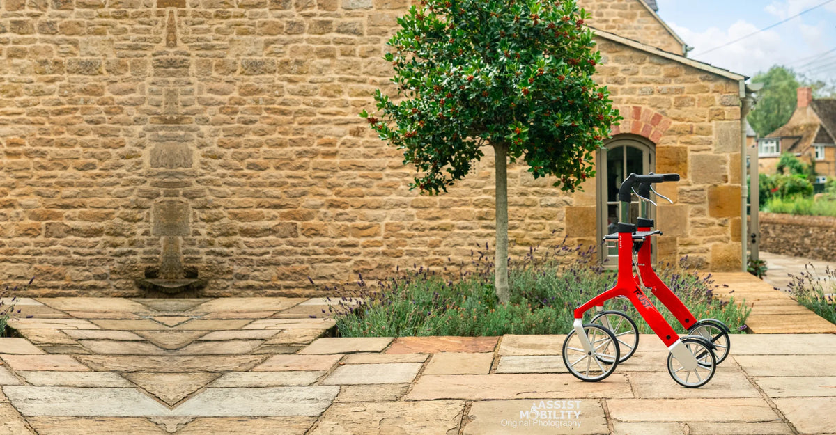 A red Trust Care Let's Fly rollator, positioned on a stone patio in front of a stone cottage with a green tree in the Cotswolds.