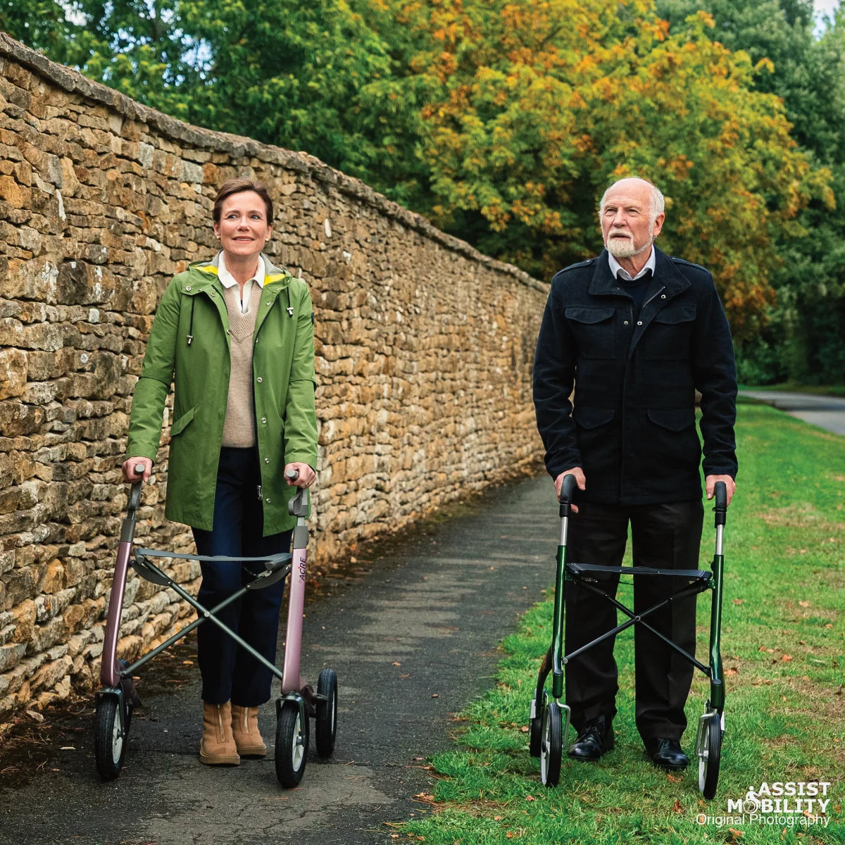 Older couple standing together with a Rose Gold byACRE Carbon Overland rollator and a British Racing Green Trust Care Let's Go Out rollator beside autumn trees