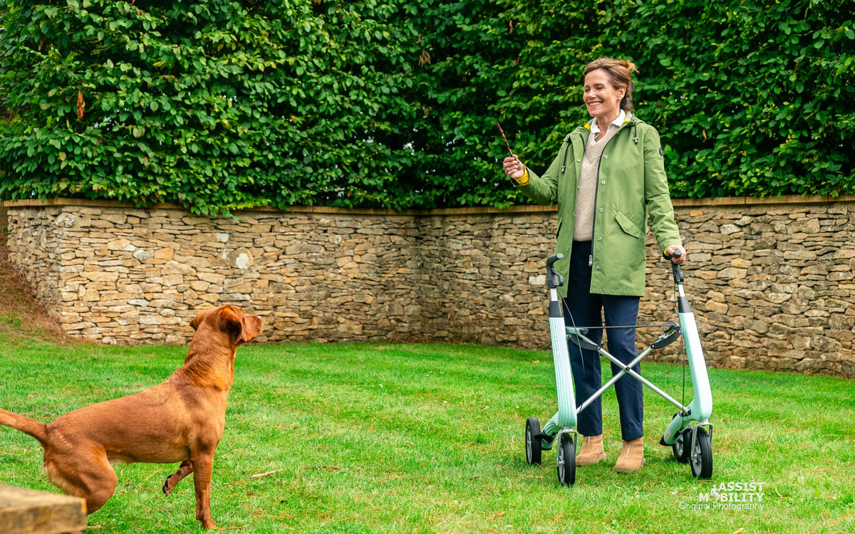 Smiling woman with a Glacier Green byACRE Nordic Pioneer rollator in a garden, with her dog on the grass