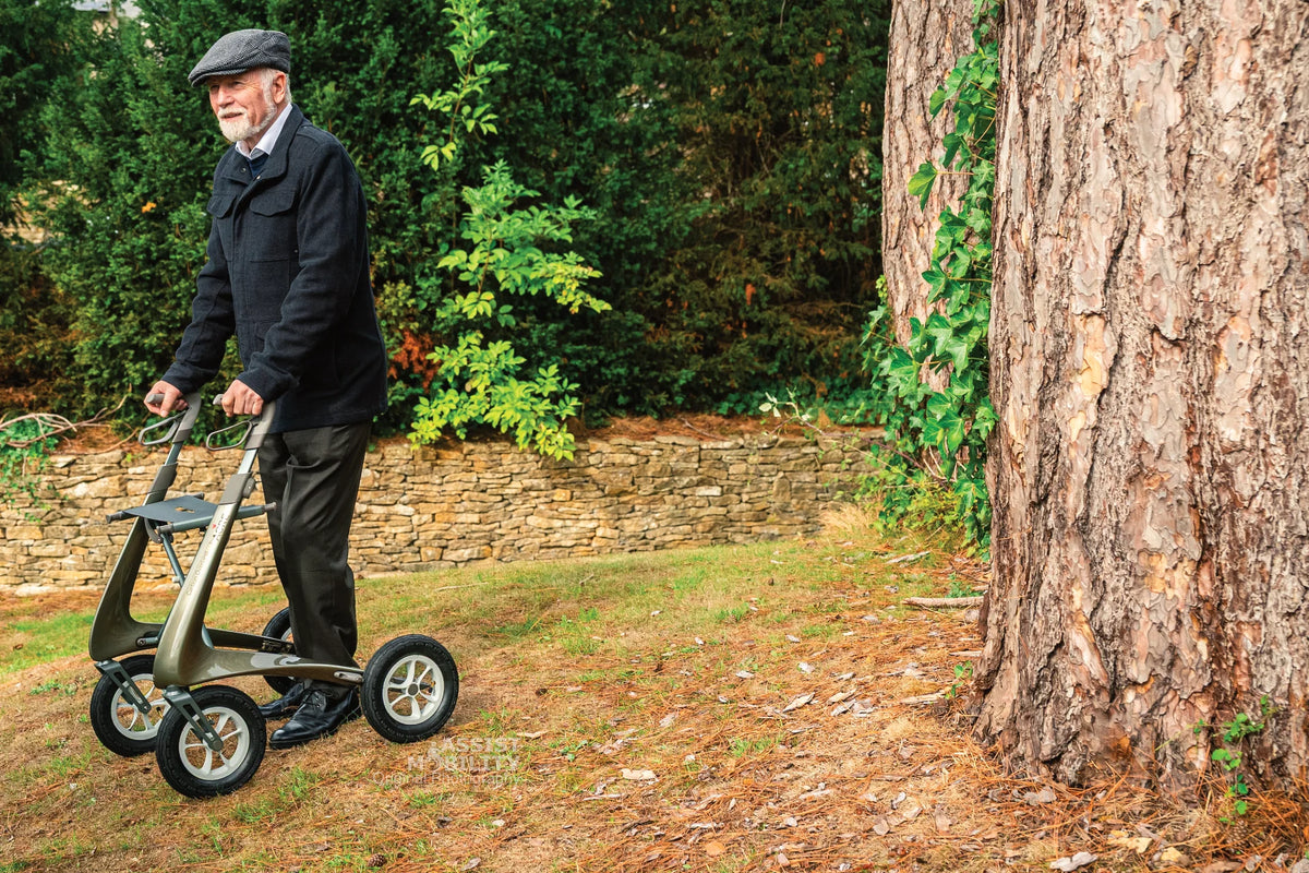 Older man in a flat cap walking across rough grass and pine needles with a byACRE Carbon Overland off-road rollator beside a large tree