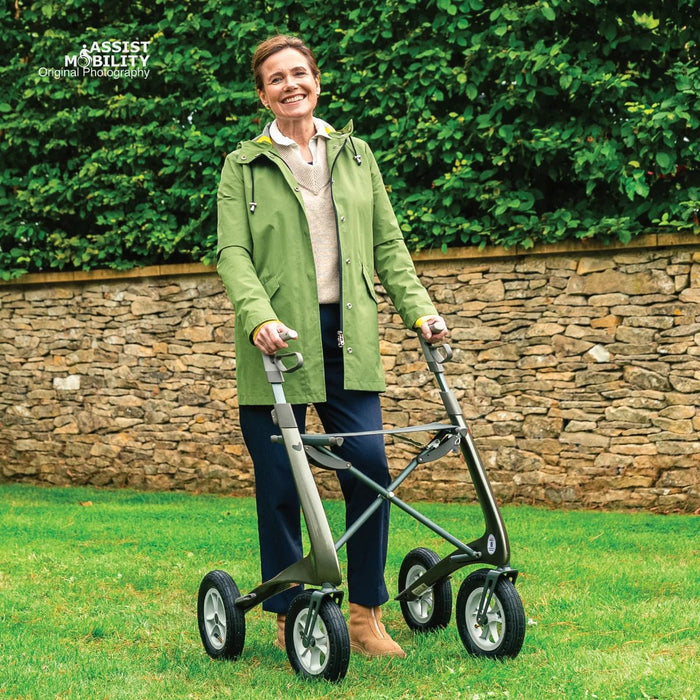 Smiling woman standing on a garden lawn with a green byACRE Carbon Overland off-road rollator in front of a stone wall