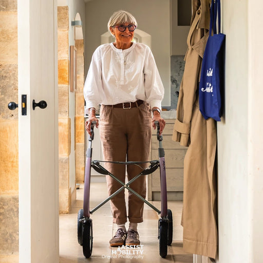 An elderly woman with short grey hair and glasses uses a byACRE Carbon Overland rollator, in a brightly lit hallway with cream walls and Cotswolds stone accents.