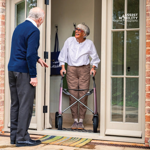 An elderly woman with short grey hair and glasses uses a byACRE Carbon Overland rollator while standing in a doorway, facing an elderly man in a blue sweater. The photo is taken from a perspective angle outside a home with brick walls and glass doors.