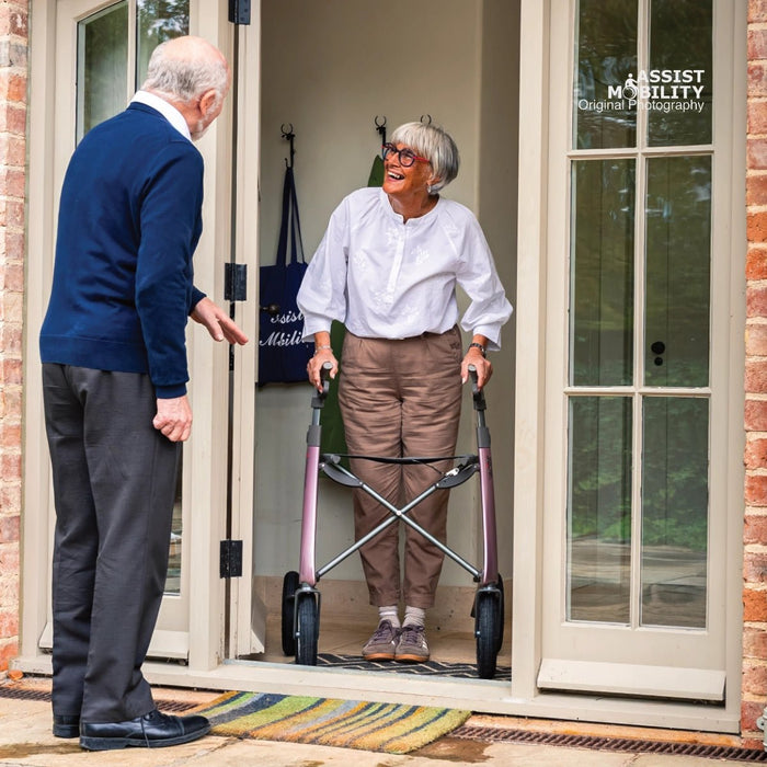 An elderly woman with short grey hair and glasses uses a byACRE Carbon Overland rollator while standing in a doorway, facing an elderly man in a blue sweater. The photo is taken from a perspective angle outside a home with brick walls and glass doors.