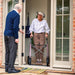 An elderly woman with short grey hair and glasses uses a byACRE Carbon Overland rollator while standing in a doorway, facing an elderly man in a blue sweater. The photo is taken from a perspective angle outside a home with brick walls and glass doors.