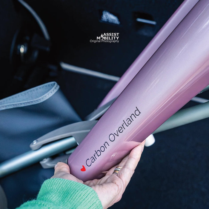 Hand holding the Rose Gold carbon fibre frame of the byACRE Carbon Overland rollator, showing the Carbon Overland logo