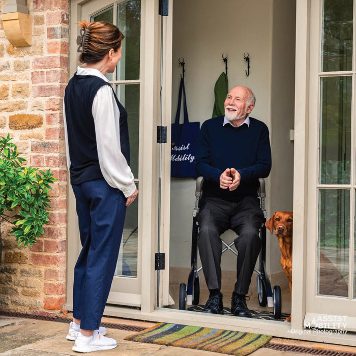 Smiling older man resting on the seat of his byACRE Carbon Ultralight rollator in his doorway, greeting a carer with his dog at his side