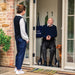 Smiling older man resting on the seat of his byACRE Carbon Ultralight rollator in his doorway, greeting a carer with his dog at his side