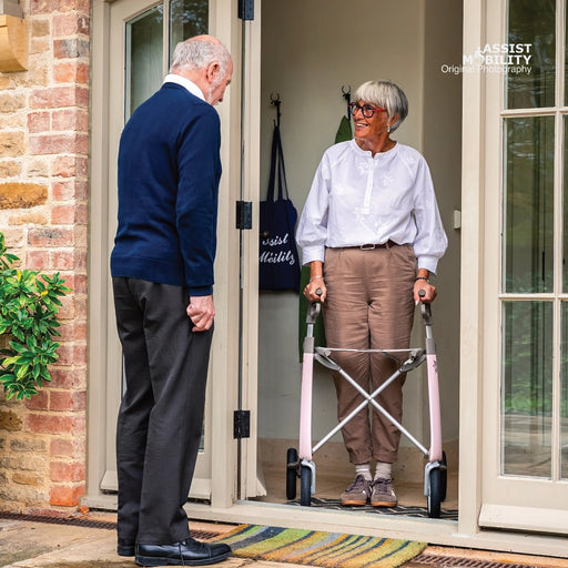 An elderly woman stands in a doorway using a pink byACRE Carbon Ultralight rollator, while an older man stands beside her outside the door. A brick wall, glass doors, and a striped rug are visible.