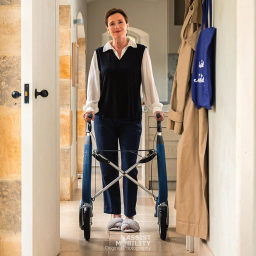 A woman using a blue byACRE Nordic Pioneer rollator in a home hallway, wearing a black vest over a white shirt and dark trousers.