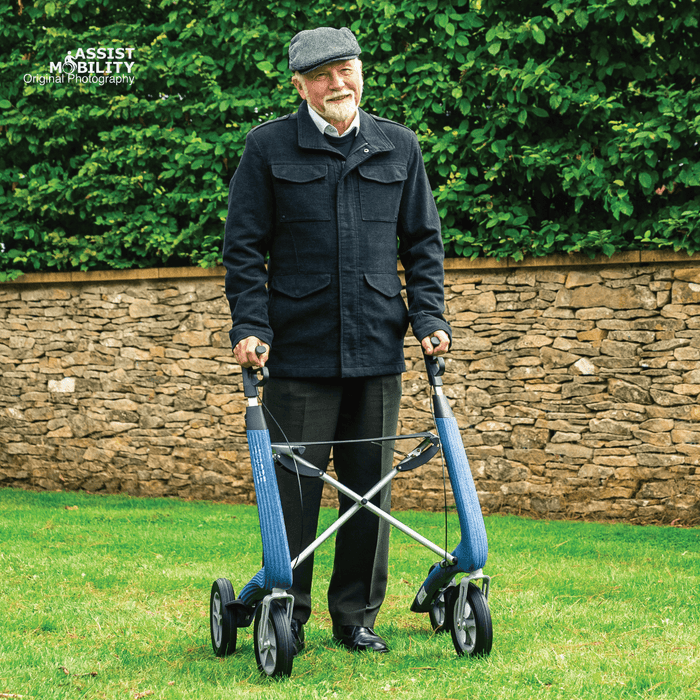 Older man in a flat cap standing on a lawn with a Baltic Blue byACRE Nordic Pioneer rollator in front of a stone wall