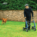 Older man walking across a lawn with a Baltic Blue byACRE Nordic Pioneer rollator while his dog runs with a stick