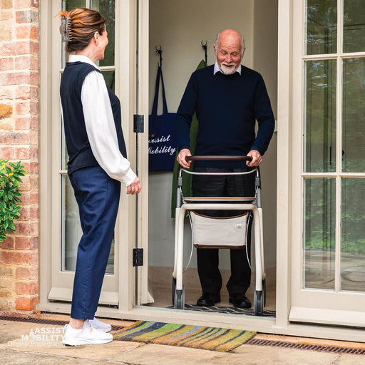 Smiling older man using a byACRE Scandinavian Butler indoor rollator with its storage bag at the open doorway of his home