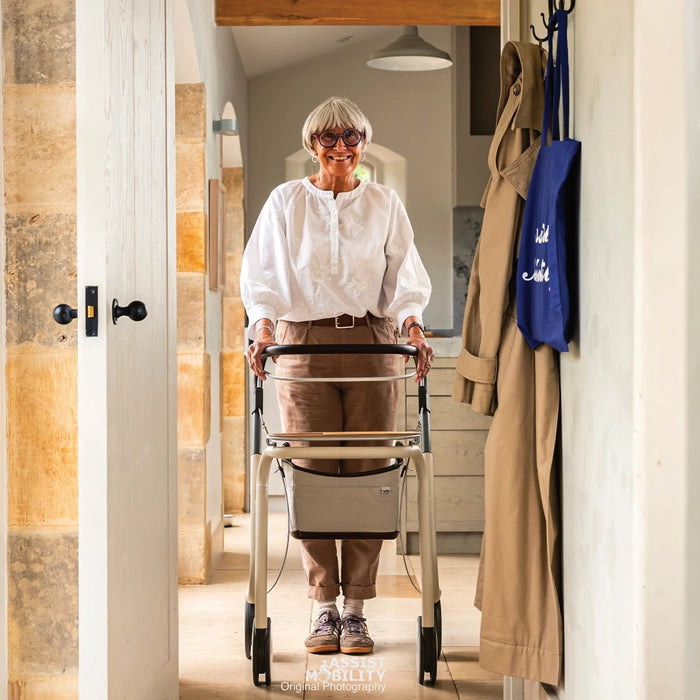 An older woman with short grey hair and glasses stands in a doorway holding a byACRE Scandinavian Butler indoor rollator with a storage basket. She is wearing a white blouse, brown trousers, and trainers.