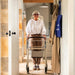 An older woman with short grey hair and glasses stands in a doorway holding a byACRE Scandinavian Butler indoor rollator with a storage basket. She is wearing a white blouse, brown trousers, and trainers.