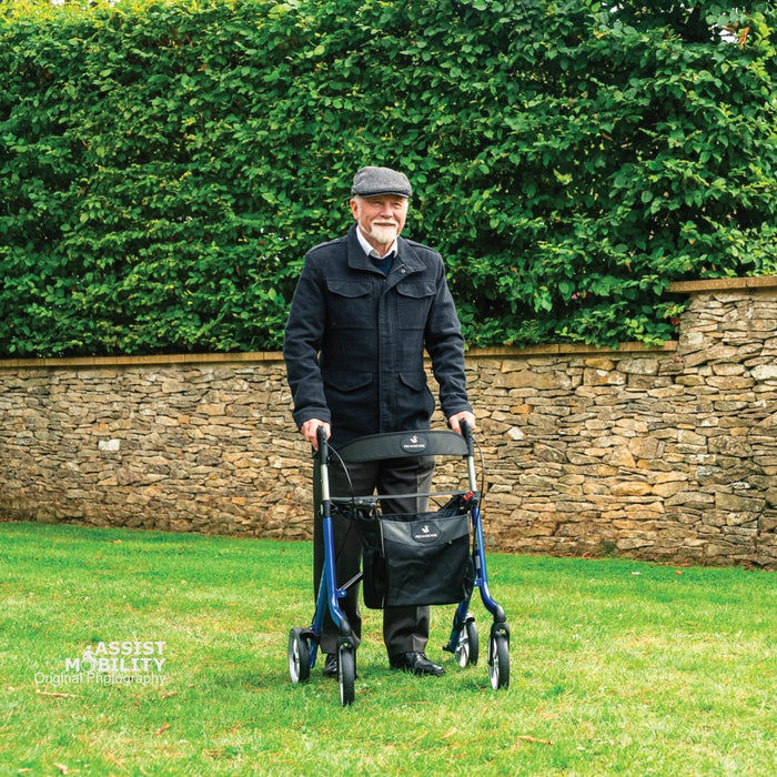 Smiling older man with an Electric Blue Rehasense Space LX rollator and storage bag on a lawn in front of a stone wall and hedge