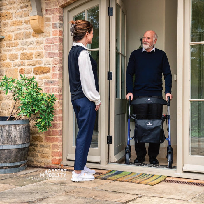 Smiling older man using an Electric Blue Rehasense Space LX rollator at the open doorway of his home as a carer arrives