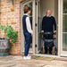 Smiling older man using an Electric Blue Rehasense Space LX rollator at the open doorway of his home as a carer arrives
