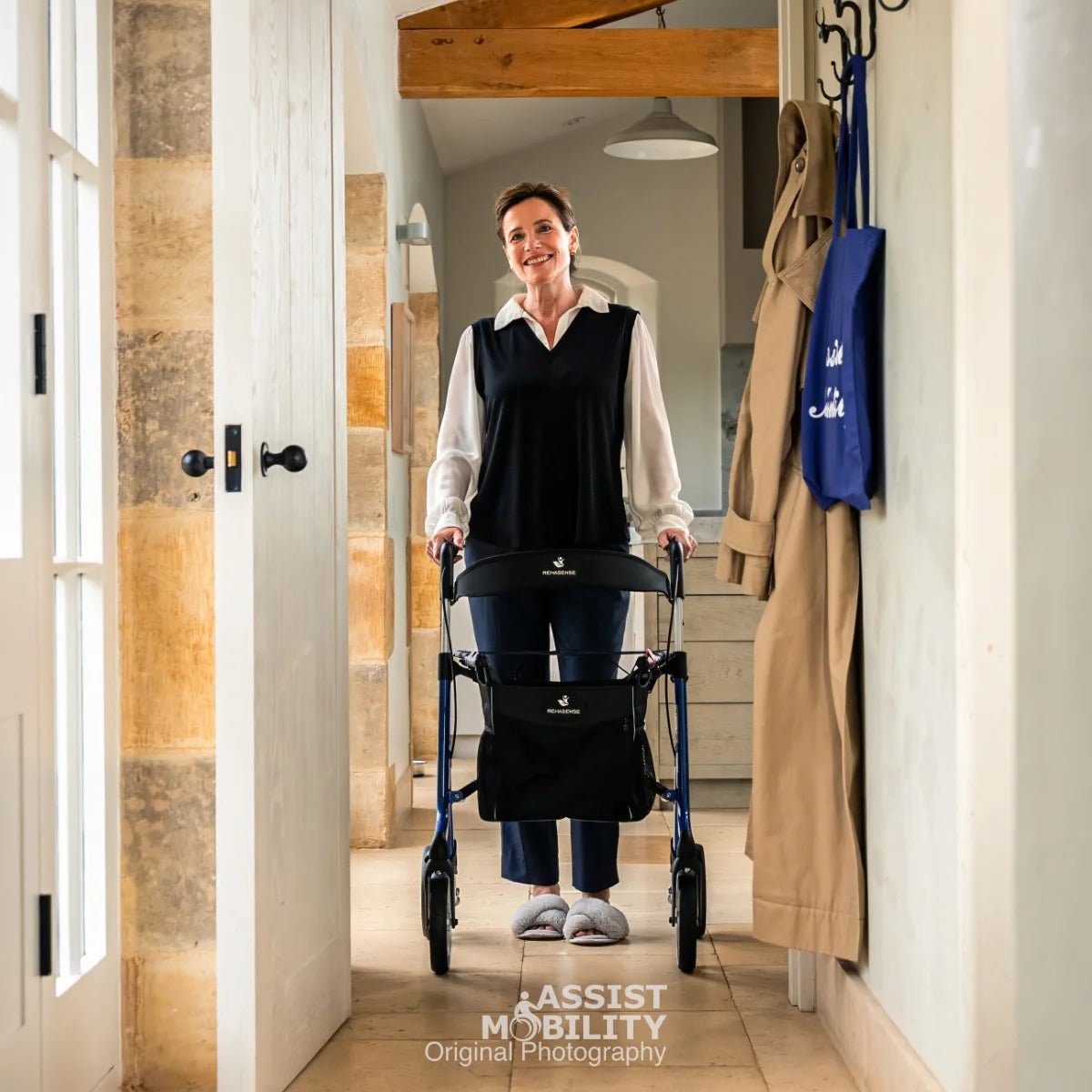 A woman pushes a blue Rehasense Space LX rollator with a shopping basket in a bright, traditional hallway with stone walls and hanging coats.