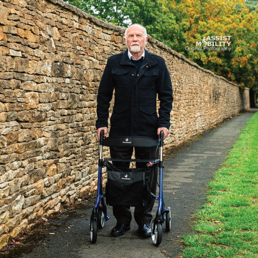 Older man standing square-on with an Electric Blue Rehasense Space LX rollator on a path between a stone wall and grass