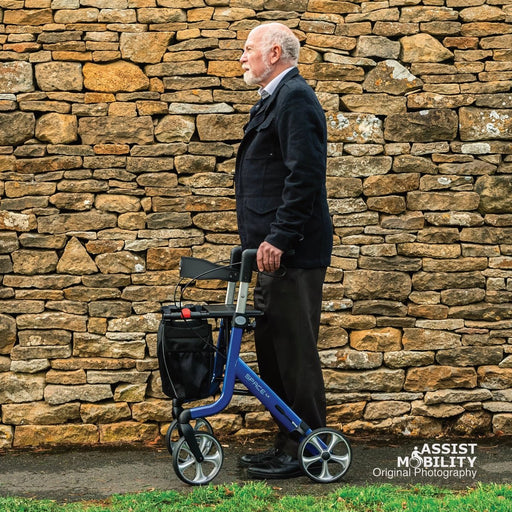 An older man in a dark jacket and trousers stands in with his Rehasense Space LX rollator in blue with a black bag, positioned in front of a rustic Cotswolds stone wall.