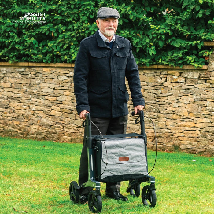 Smiling older man in a flat cap standing on a lawn with a Rollz Air Carbon Fibre rollator and its grey bag by a stone wall