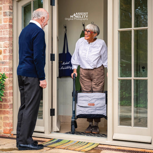 An elderly woman with short grey hair and glasses stands in a doorway using a Rollz Air Carbon Fibre rollator, while an older man in a navy sweater stands outside. The woman wears a white button-up shirt and brown trousers.