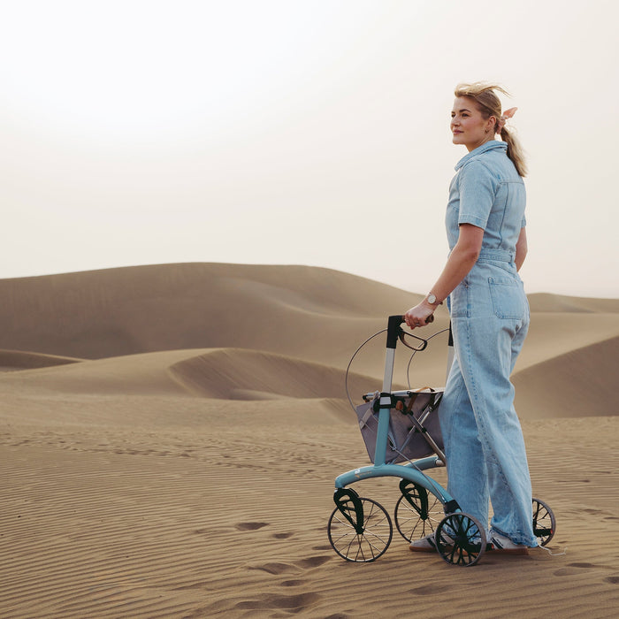 A woman in sand dunes pushes a Saljol Allround Rollator Superlight Ice Blue rollator  with a net basket. She is wearing a light blue denim boiler suit.