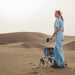 A woman in sand dunes pushes a Saljol Allround Rollator Superlight Ice Blue rollator  with a net basket. She is wearing a light blue denim boiler suit.