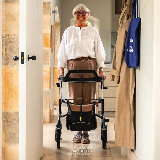 An elderly woman with short grey hair and glasses uses a Saljol Carbon Fibre rollator  in a bright hallway. She is wearing a white long-sleeve blouse, brown trousers, and trainers.