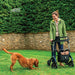 Laughing woman standing with a Saljol Carbon Fibre rollator in her garden while her dog plays on the lawn
