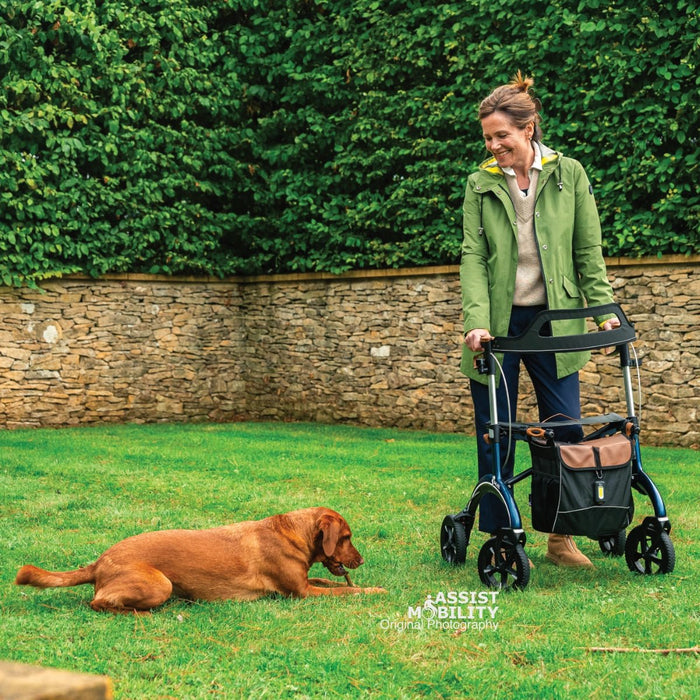 Woman with a Saljol Carbon Fibre rollator on a garden lawn, smiling at her dog lying nearby, with a stone wall and hedge behind
