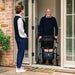 Older man at his front door with a Saljol Carbon Fibre rollator and shopping bag, greeting a carer outside a Cotswold stone house