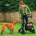 Smiling woman leaning on her Saljol Carbon Fibre rollator to give her dog a treat on a garden lawn