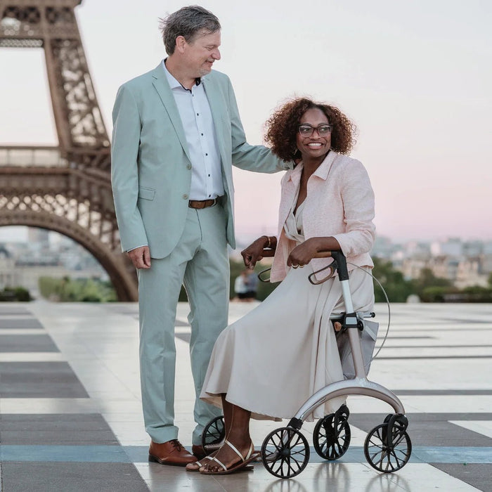 Smiling woman sitting on the seat of a Saljol Carbon Superlight rollator beside her partner, with the Eiffel Tower behind them