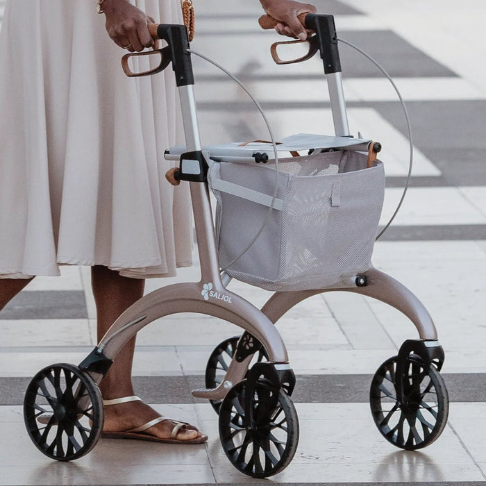 Close-up of a woman walking with a Rose Gold Saljol Carbon Superlight rollator, showing its curved frame, mesh bag and large wheels