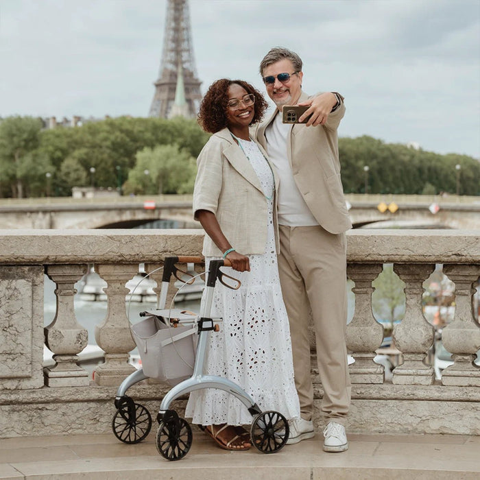 Smiling couple taking a selfie on a Paris bridge with the Eiffel Tower behind, a Saljol Carbon Superlight rollator beside them