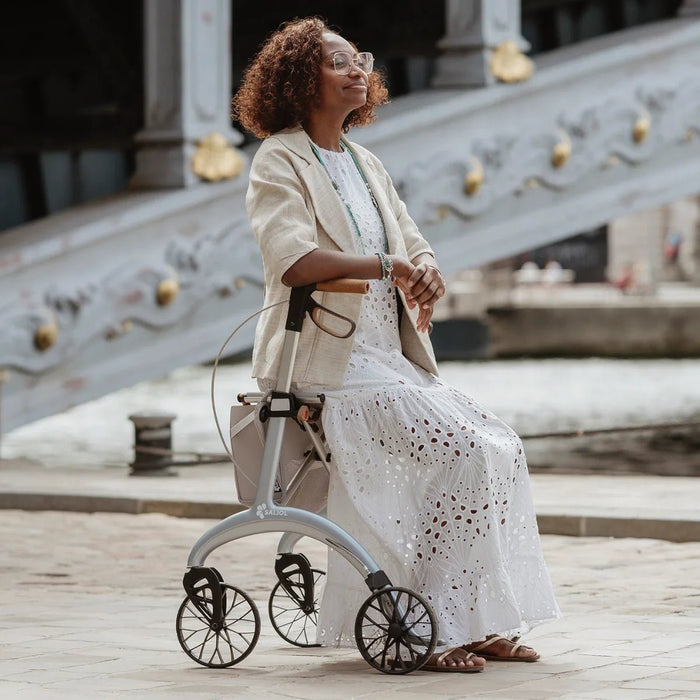 Woman resting on the seat of a Saljol Carbon Superlight rollator beside an ornate Paris bridge on a sunny day
