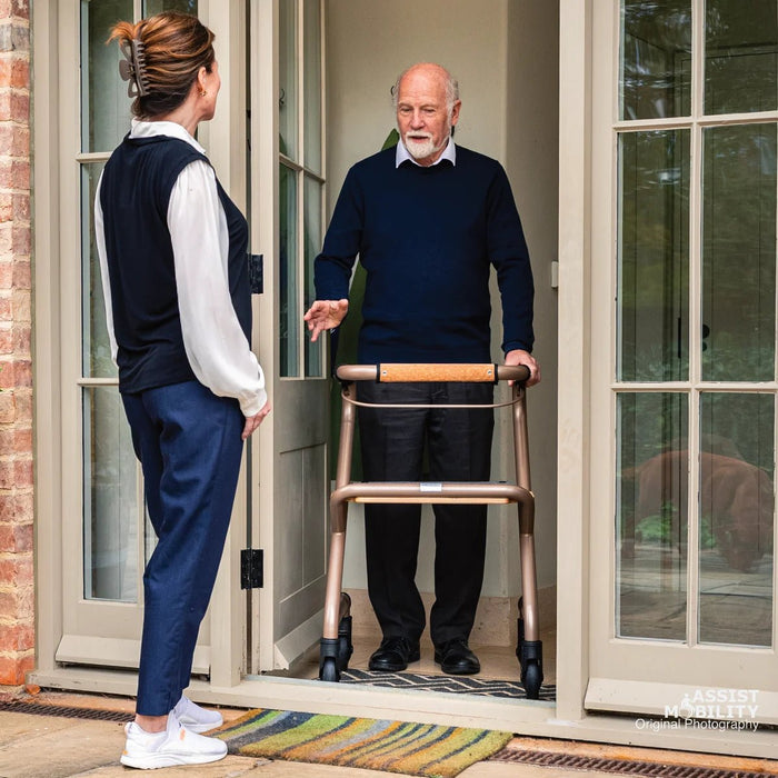 Smiling older man standing in his doorway with a slim Saljol indoor rollator, chatting with a carer on the doorstep