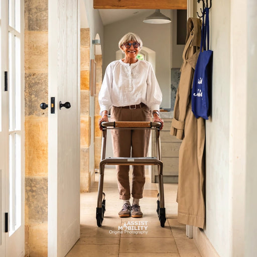 An elderly woman with short grey hair and glasses uses a Saljol Page indoor rollator  in a bright hallway. She is wearing a white long-sleeve blouse, brown trousers, and trainers.