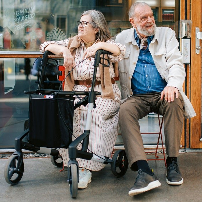 An elderly couple seated next to the Troja Original Black Series rollator. 
