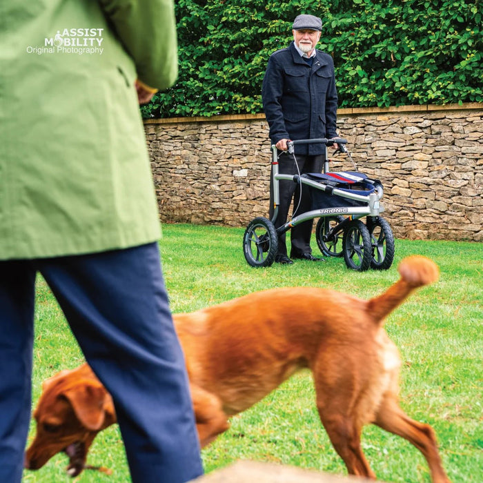 Older man with a Trionic Veloped Sport off-road rollator on a garden lawn, with a woman and their dog in the foreground
