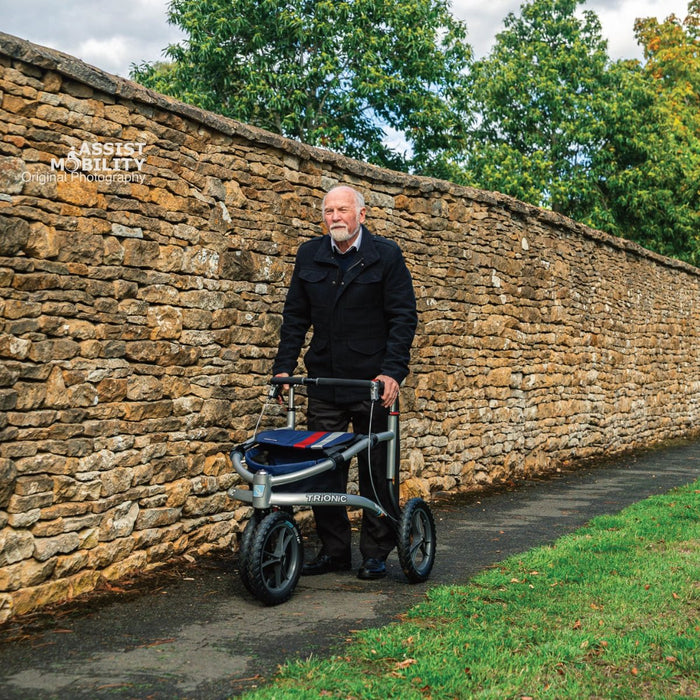 Older man standing with the Trionic Veloped Sport off-road rollator on a path beside a Cotswold stone wall