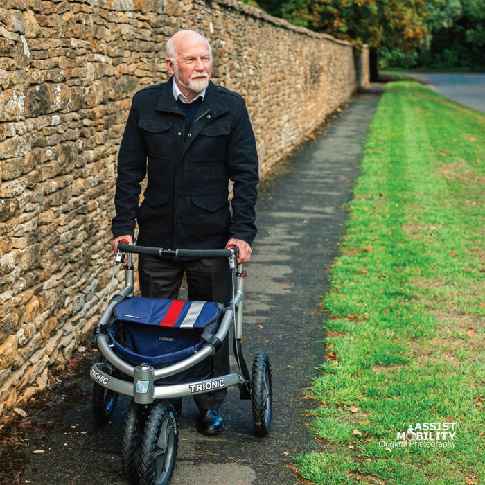 Older man walking with the Trionic Veloped Sport off-road rollator along a village path beside a stone wall and grass verge
