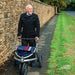 Older man walking with the Trionic Veloped Sport off-road rollator along a village path beside a stone wall and grass verge