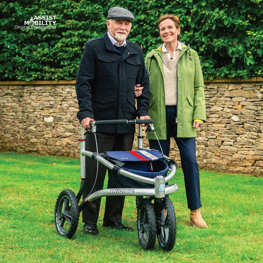 Smiling couple standing arm in arm on a garden lawn, the man with a Trionic Veloped Sport off-road rollator