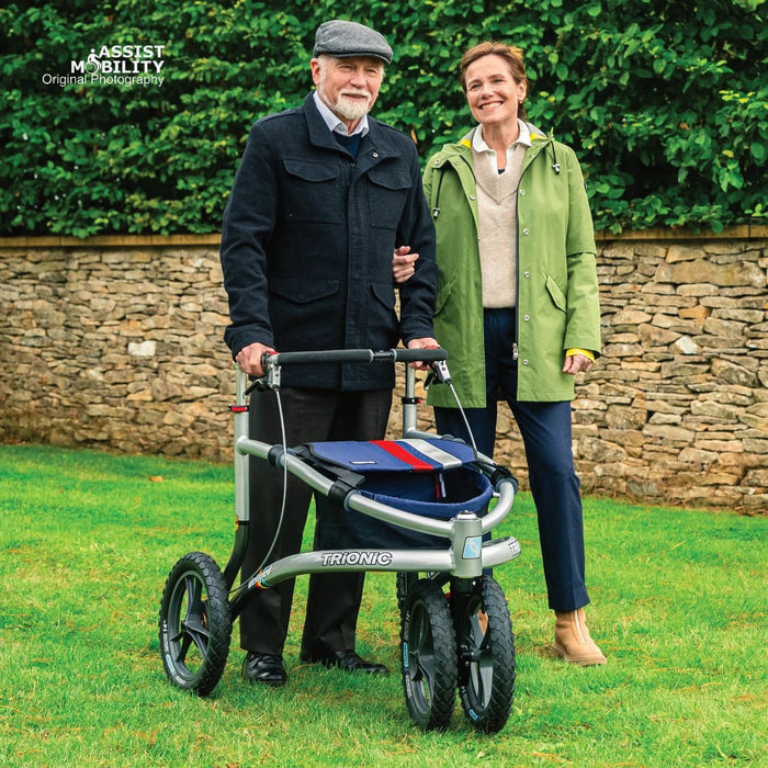 Smiling couple standing arm in arm on a garden lawn, the man with a Trionic Veloped Sport off-road rollator