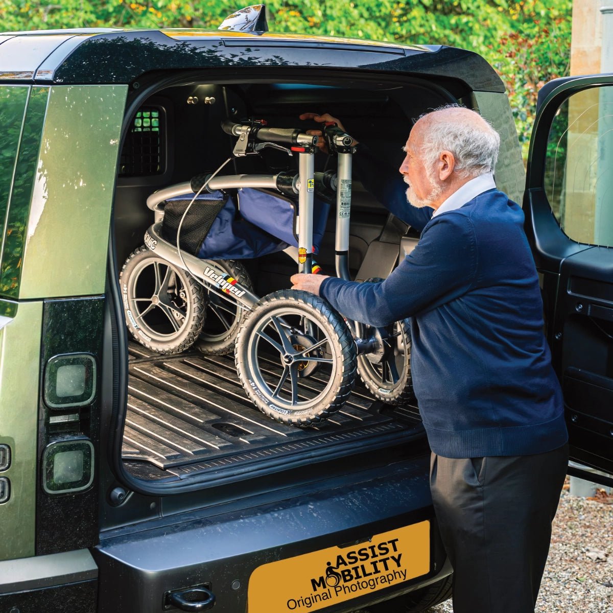 An older man in a navy sweater and dark trousers is loading his Trionic Veloped Sport Off Road Rollator into the boot of a car.