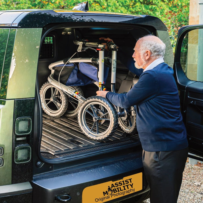 An older man in a navy sweater and dark trousers is loading his Trionic Veloped Sport Off Road Rollator into the boot of a car.