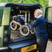 An older man in a navy sweater and dark trousers is loading his Trionic Veloped Sport Off Road Rollator into the boot of a car.